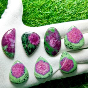 Pu&ograve; includere: Sette cabochon lucidati di rubino in zoisite, ognuno caratterizzato da un vibrante centro magenta-rosa circondato da una matrice minerale verde e nera maculata. Le pietre sono modellate in varie forme, tra cui gocce, ovali e una mezzaluna.