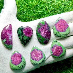Pu&ograve; includere: Sette gemme levigate di rubino in zoisite con vivaci centri magenta-rosa circondati da inclusioni minerali screziate verde lime e nere. Le pietre sono modellate in varie forme a goccia, ovali e a mezzaluna con superfici lisce.