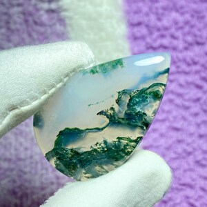 May include: A polished, teardrop-shaped gemstone with a translucent white base. The stone features intricate patterns of green moss-like inclusions, creating a natural, artistic design. The gem is held by a gloved hand.
