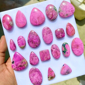 May include: A collection of polished, vibrant pink gemstones in various shapes, including ovals, teardrops, and squares. The stones have unique patterns and textures, with some showing hints of green and brown. The gemstones are displayed on a white surface.