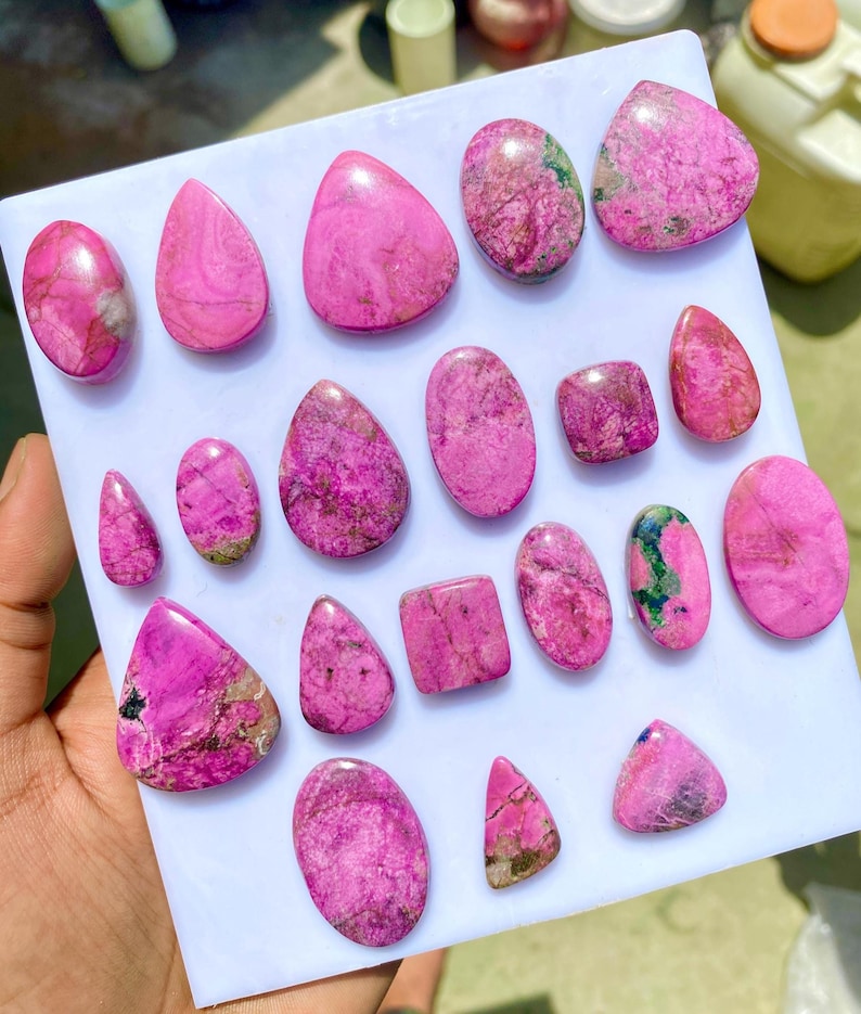 May include: A collection of polished, vibrant pink gemstones in various shapes, including ovals, teardrops, and squares. The stones have unique patterns and textures, with some showing hints of green and brown. The gemstones are displayed on a white surface.