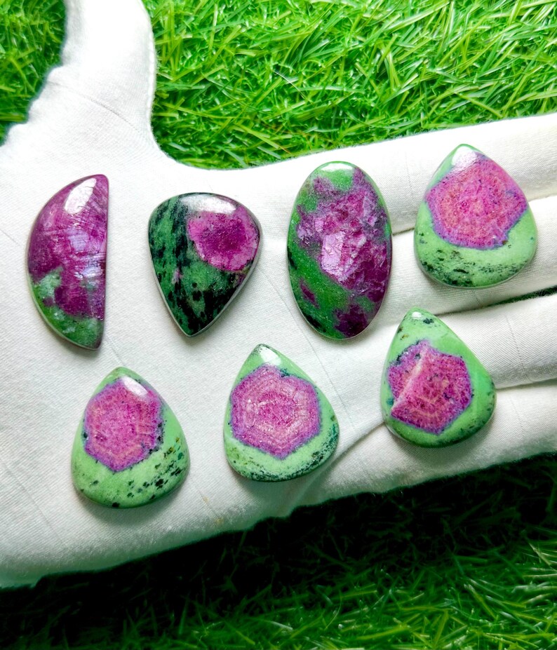 Pu&ograve; includere: Sette cabochon lucidati di zoisite con inclusioni di rubino, caratterizzati da un mix di forme a goccia, ovali e semicircolari. Ogni pietra presenta un vivace centro rosa magenta circondato da una matrice minerale verde chiaro e nera.