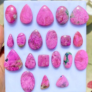 May include: A collection of polished pink gemstones in various shapes, including oval, teardrop, and triangular forms. The stones display a marbled texture with darker pink and brown inclusions, arranged on a white surface.