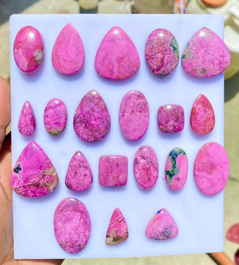 May include: A collection of polished pink gemstones in various shapes, including oval, teardrop, and triangular forms. The stones display a marbled texture with darker pink and brown inclusions, arranged on a white surface.