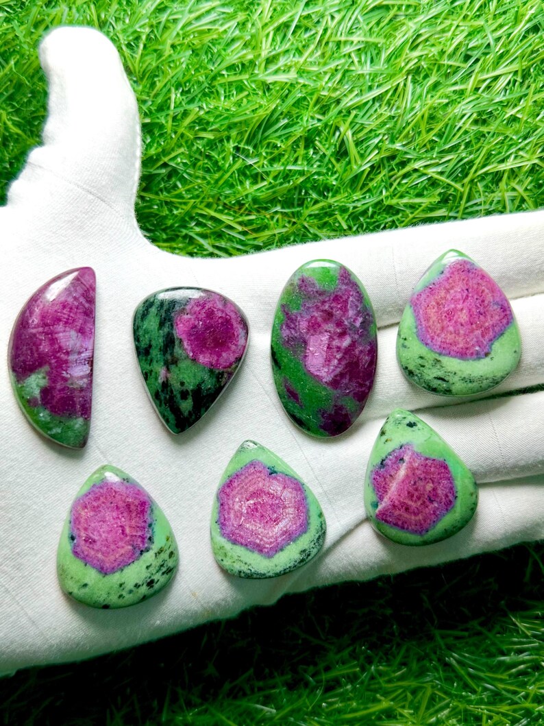 Pu&ograve; includere: Sette cabochon lucidati di rubino in zoisite, ognuno caratterizzato da un vibrante centro magenta-rosa circondato da una matrice minerale verde e nera maculata. Le pietre sono modellate in varie forme, tra cui gocce, ovali e una mezzaluna.
