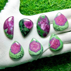 Pu&ograve; includere: Sette cabochon lucidati di zoisite con inclusioni di rubino, caratterizzati da un mix di forme a goccia, ovali e semicircolari. Ogni pietra presenta un vivace centro rosa magenta circondato da una matrice minerale verde chiaro e nera.