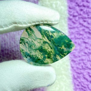 May include: A teardrop-shaped gemstone with a polished surface. The stone features a swirling pattern of green and white, resembling a landscape. The gemstone is held by a gloved hand, showcasing its natural beauty and unique design.