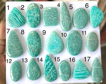 Può includere: Diciassette cabochon di amazzonite lucidati disposti su un pannello espositivo bianco, numerati da 1 a 17. Ogni pietra presenta un distinto colore verde-azzurro con inclusioni bianche, fibrose e venate, in varie forme come goccia, ovale e rettangolo.
