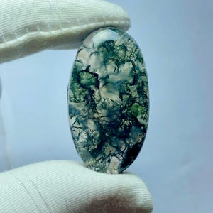 May include: An oval-shaped gemstone with a moss agate pattern. The stone features a translucent base with intricate green and white inclusions, resembling a miniature landscape. The gemstone is held by a gloved hand.