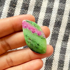 以下が含まれることがあります： 鮮やかな色の組み合わせが特徴的な、磨かれた宝石。緑、ピンク、黒の色合いが印象的です。ピンクの部分は中央にあり、緑と黒の色は石全体に分布しています。宝石は手で持たれています。