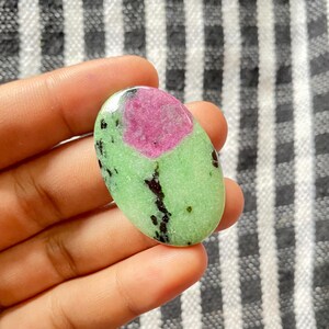 以下が含まれることがあります： 鮮やかなピンク色の中心部と明るい緑色の外側を持つ楕円形の宝石。緑色の部分には黒い斑点と静脈があります。宝石は、白と黒のストライプの背景に対して、手で持たれています。