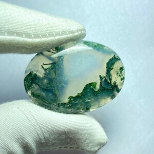 May include: Oval-shaped moss agate gemstone. The translucent stone features a landscape of green, blue, and white patterns, resembling a miniature natural scene. The stone is held by a gloved hand.