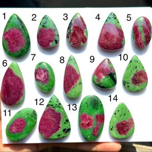 Op de afbeelding: Een collectie van veertien gepolijste zoisiet edelstenen met robijninsluitingen, genummerd van één tot veertien. De stenen hebben levendige magenta-rode vlekken tegen een heldergroene achtergrond, in verschillende druppel- en ovale vormen.