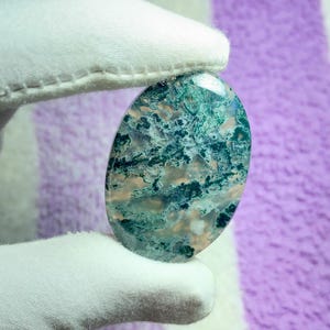 May include: An oval-shaped gemstone with a unique pattern of green, blue, and beige hues. The stone is held by a gloved hand, showcasing its polished surface and natural variations. The gemstone's colors and patterns create a striking visual effect.