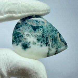 May include: A polished, teardrop-shaped gemstone with a white base and intricate patterns of green and dark teal. The stone is held by a white gloved hand, showcasing its natural beauty and unique veining.