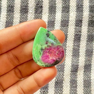 以下が含まれることがあります： 鮮やかな緑色の外観と深い赤色の内観を持つ、涙の形をした宝石。石は研磨された表面を持ち、色とテクスチャの自然なパターンを示しています。 宝石は手で持たれており、背景はストライプです。