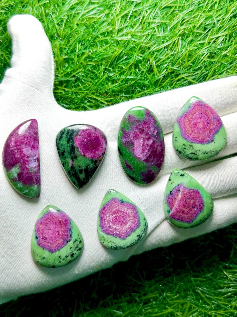 Pu&ograve; includere: Sette gemme levigate di rubino in zoisite con vivaci centri magenta-rosa circondati da inclusioni minerali screziate verde lime e nere. Le pietre sono modellate in varie forme a goccia, ovali e a mezzaluna con superfici lisce.