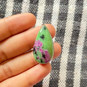 以下が含まれることがあります： 鮮やかな緑色のベースにピンクと黒のインクルージョンが散りばめられた、涙型の宝石。 研磨された石が持ち上げられ、その自然な模様と色が示されています。 この宝石は、ジュエリーやその他の工芸品に使用できます。
