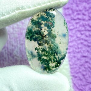 May include: An oval-shaped moss agate gemstone. The translucent stone features green, tree-like inclusions, with hints of blue and red. The stone is held by a gloved hand against a purple background. This gemstone is ideal for jewelry making.