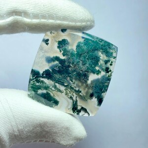 May include: A square-cut gemstone with a translucent base, featuring intricate patterns of green and white, resembling a miniature landscape. The stone is held by a gloved hand, showcasing its polished surface and natural beauty.