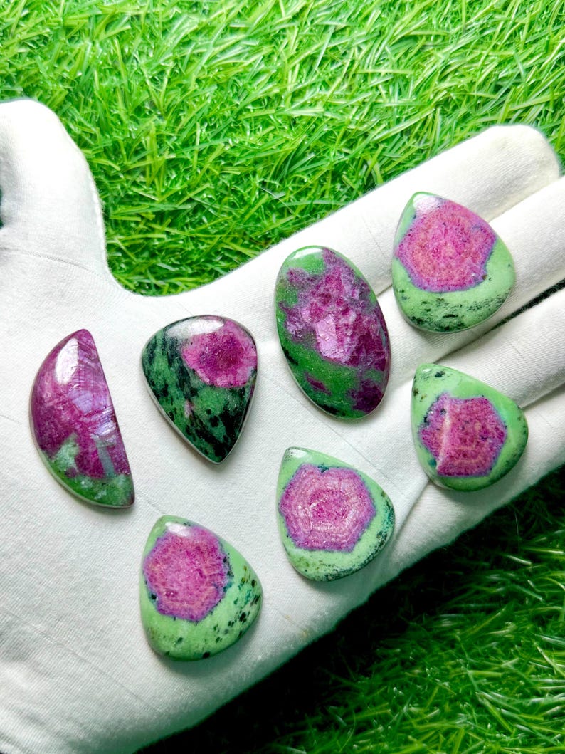 Pu&ograve; includere: Sette cabochon levigati di zoisite con inclusioni di rubino, caratterizzati da vivaci centri magenta circondati da pietra verde e nera maculata. Le pietre sono modellate in varie forme a goccia, ovali e semicircolari con superfici lisce e lucide.
