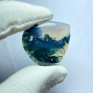 May include: A translucent, polished gemstone with a landscape-like pattern. The stone features shades of blue, green, and white, resembling a miniature natural scene. The gemstone is held by a gloved hand.