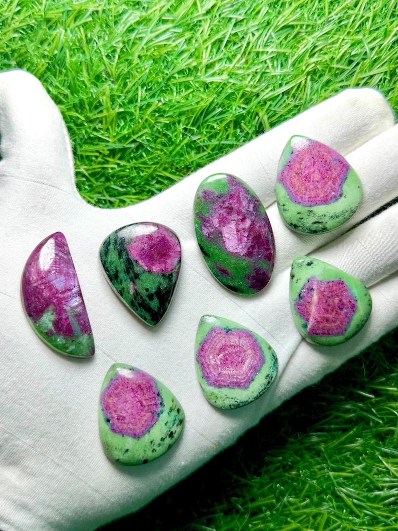 Pu&ograve; includere: Sette cabochon levigati di zoisite con inclusioni di rubino, caratterizzati da vivaci centri magenta circondati da pietra verde e nera maculata. Le pietre sono modellate in varie forme a goccia, ovali e semicircolari con superfici lisce e lucide.