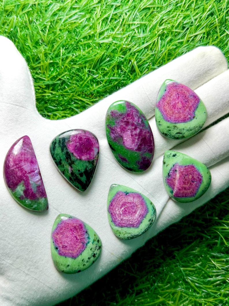 Pu&ograve; includere: Sette cabochon levigati di zoisite con inclusioni di rubino, ognuno dei quali presenta un vivace centro magenta-rosa circondato da motivi minerali screziati verde lime e neri. Le pietre sono disposte in varie forme, tra cui gocce e ovali.