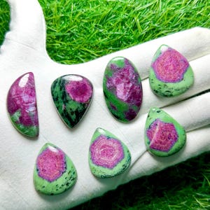 Pu&ograve; includere: Sette pietre preziose di zoisite lucida con centri magenta vibranti circondati da inclusioni minerali verdi e nere screziate. Le pietre sono modellate in varie forme a goccia, ovali e a mezzaluna con superfici lisce e riflettenti.