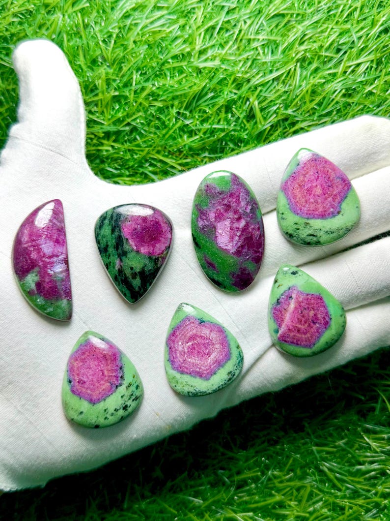 Pu&ograve; includere: Sette pietre preziose di zoisite lucida con centri magenta vibranti circondati da inclusioni minerali verdi e nere screziate. Le pietre sono modellate in varie forme a goccia, ovali e a mezzaluna con superfici lisce e riflettenti.