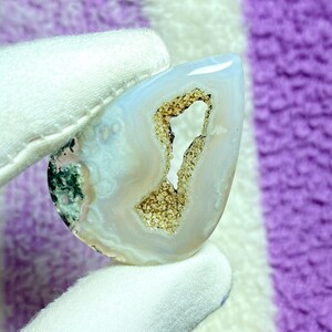 May include: A polished, teardrop-shaped gemstone with a white and light blue exterior. The center features a cavity filled with a sparkling, golden-brown crystalline structure. The gemstone is held by a gloved hand.