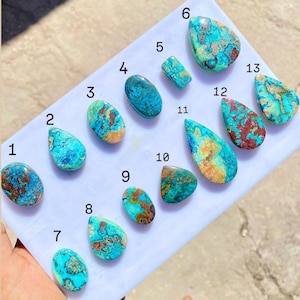 May include: A collection of thirteen polished gemstones, each with unique patterns and colors. The stones feature a mix of turquoise, brown, and gold hues, with varying shapes including ovals, teardrops, and rectangles. The gemstones are displayed on a white surface.