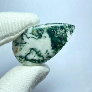 May include: A polished, teardrop-shaped gemstone with a white base and intricate green moss-like inclusions. The stone is held by a gloved hand, showcasing its natural patterns and translucent quality. The gemstone's surface is smooth and reflective.