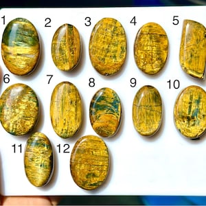 May include: Twelve polished oval and shield-shaped gemstones arranged on a white surface, numbered one through twelve. Each stone features a chatoyant pattern with golden-yellow and deep green streaks, displaying a smooth, reflective surface texture.