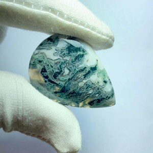 May include: A polished gemstone with a teardrop shape. The stone features a swirling pattern of green and white, with hints of yellow. The gemstone is held between two white gloved hands.