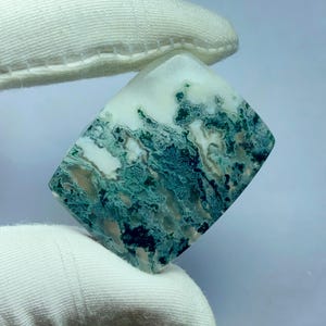 May include: A polished, square-cut gemstone with a white and teal-green pattern. The stone is held by a gloved hand, showcasing the intricate, natural design. The gemstone's surface has a marbled appearance, with swirling colors.