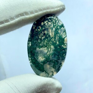 May include: An oval-shaped gemstone with a translucent base and intricate green moss-like inclusions. The stone is held between two white gloved fingers, showcasing its natural patterns and colors. The gemstone's polished surface reflects light, highlighting its unique internal structures.