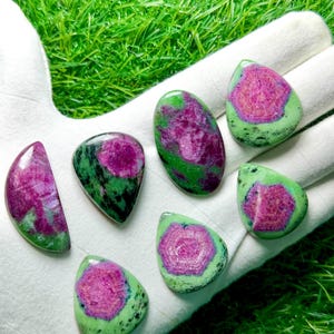 Pu&ograve; includere: Sette cabochon levigati di zoisite con inclusioni di rubino, ognuno dei quali presenta un vivace centro magenta-rosa circondato da motivi minerali screziati verde lime e neri. Le pietre sono disposte in varie forme, tra cui gocce e ovali.