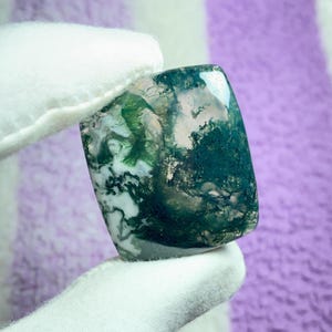May include: A polished, rectangular moss agate gemstone. The stone features a translucent white base with intricate patterns of green and dark teal, resembling a miniature landscape. The stone is held by a gloved hand.