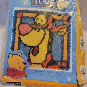 May include: A Tigger latch hook kit in plastic packaging featuring a yellow, orange, and blue yarn design of the character. The packaging includes the text Pooh, Tigger Latch Hook Kit, and Caron International. The kit is 12 inches by 12 inches.