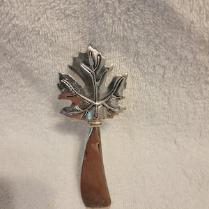 May include: A silver-toned metal spreader featuring a decorative handle shaped like a maple leaf. The leaf design includes detailed embossed veins and a textured surface, while the blade is smooth, polished, and curved for spreading food items.