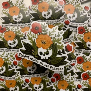 May include: A white sticker with a skull surrounded by orange and red flowers and green leaves. The sticker has the text "Bloom where you are planted" written on it.