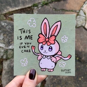 May include: A pink bunny with a red bow and pink wings on a green background. The text "This is me if you even care" is written in black. The image is signed "Poptort Studio".