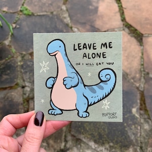 May include: A light green card with a cartoon blue dinosaur with a pink belly. The dinosaur is standing with its arms crossed and its tail curled. The text on the card reads "Leave me alone or I will eat you". The card is signed "Poptort Studio".