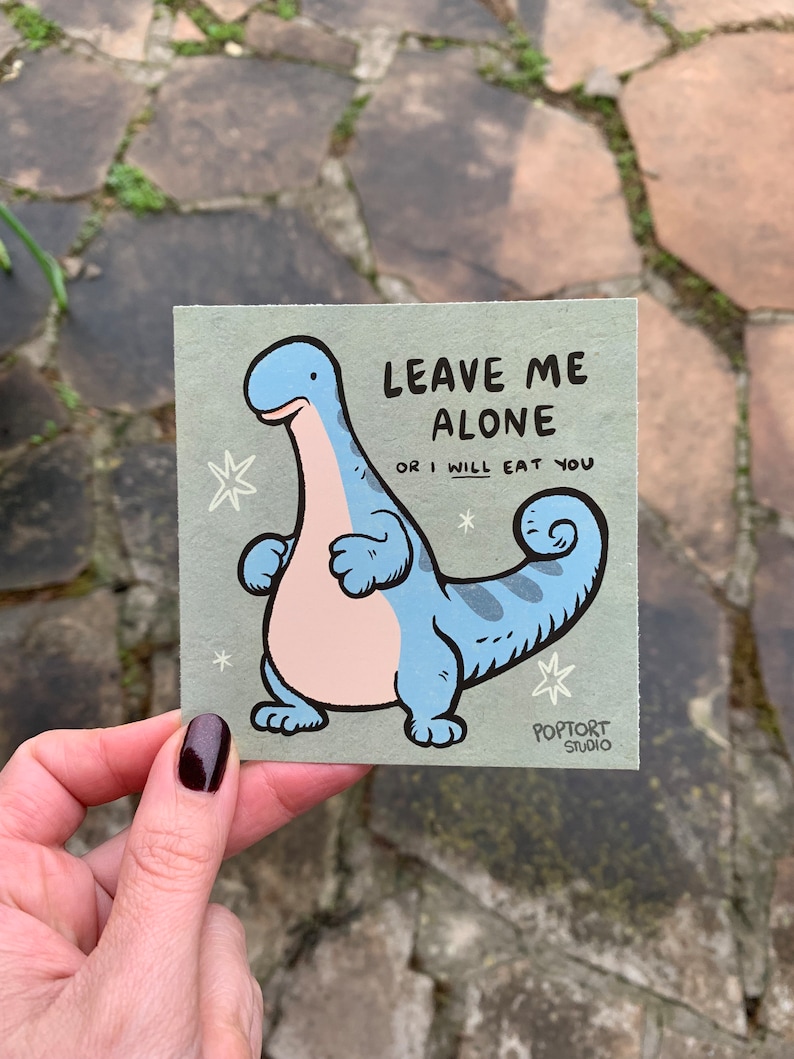 May include: A light green card with a cartoon blue dinosaur with a pink belly. The dinosaur is standing with its arms crossed and its tail curled. The text on the card reads "Leave me alone or I will eat you". The card is signed "Poptort Studio".
