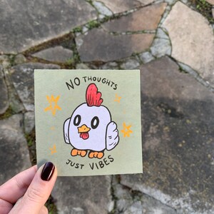 May include: A green card with a white chicken illustration and the text "NO THOUGHTS JUST VIBES". The chicken has red comb and wattles, orange feet, and black eyes. There are yellow stars around the chicken.