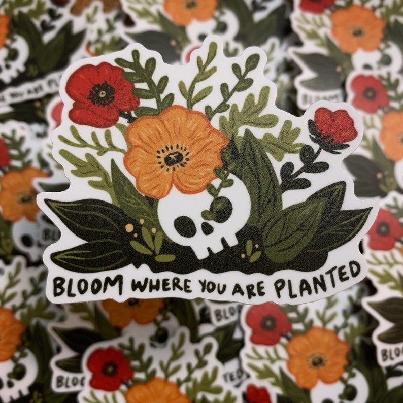 May include: A white sticker with a skull surrounded by orange and red flowers and green leaves. The text "Bloom where you are planted" is written in a black font at the bottom of the sticker.