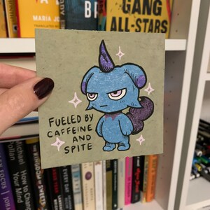 May include: A blue and purple cartoon creature with a single horn on its head. The creature is standing on a light brown background with the text "Fueled by Caffeine and Spite" written below it.