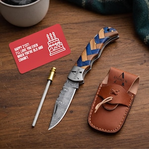 May include: A personalized pocket knife with a patterned wooden handle and a leather sheath, next to a sharpening rod and a red card that says "HAPPY 21ST! I'LL LOVE YOU EVEN WHEN YOU'RE OLD AND CRANKY."