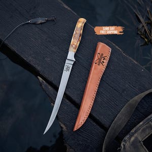 May include: A silver-colored fillet knife with a light brown patterned handle and a matching tan leather sheath. The blade features a small logo with crossed fishing rods and the text Rivermaster. A fishing lure is positioned to the left.